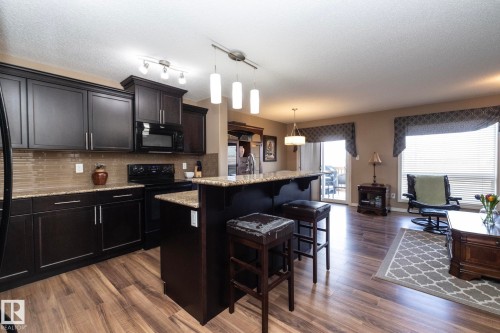 The open concept living area features wood-look flooring, a kitchen with dark cabinetry, granite countertops, and a tiled backsplash, and a living space with large windows providing natural light - 130 Kirpatrick Way, Leduc, AB - Indoor Photo Showing Kitchen