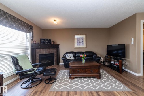 The living area features wood-look flooring, a fireplace with a tile surround and dark wood mantel, and a large window with blinds - 130 Kirpatrick Way, Leduc, AB - Indoor Photo Showing Living Room With Fireplace