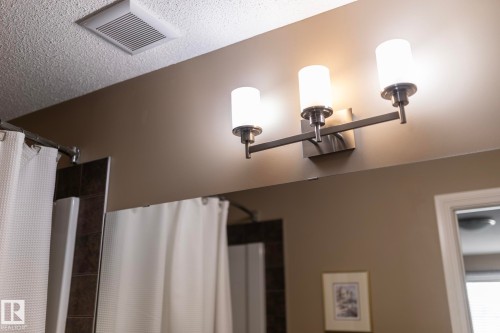 Bathroom featuring a contemporary three-light vanity fixture with brushed metal accents and frosted glass shades, a textured ceiling, and an exhaust fan - 130 Kirpatrick Way, Leduc, AB - Indoor