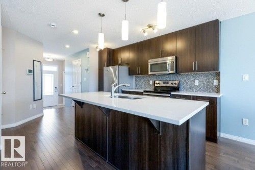 The kitchen features dark wood cabinetry, stainless steel appliances, a white island countertop with an integrated sink, and a mosaic tile backsplash - 3119 Carpenter Landing Landing, Edmonton, AB - Indoor Photo Showing Kitchen With Stainless Steel Kitchen With Double Sink With Upgraded Kitchen