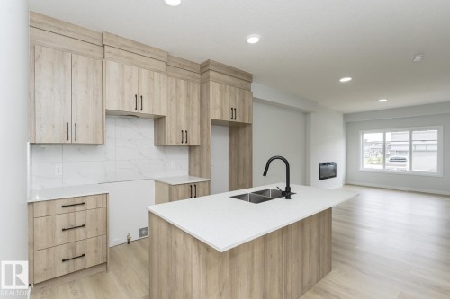 The kitchen features light wood cabinetry, a white countertop island with a double sink and a black faucet, and a tiled backsplash - 6371 King Wynd, Edmonton, AB - Indoor Photo Showing Kitchen With Double Sink
