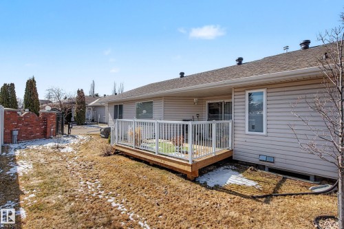 The property features a light-colored siding exterior, a brown shingled roof, and a wooden deck with white railings - 21 1850 Mill Woods Road E, Edmonton, AB - Outdoor With Deck Patio Veranda With Exterior