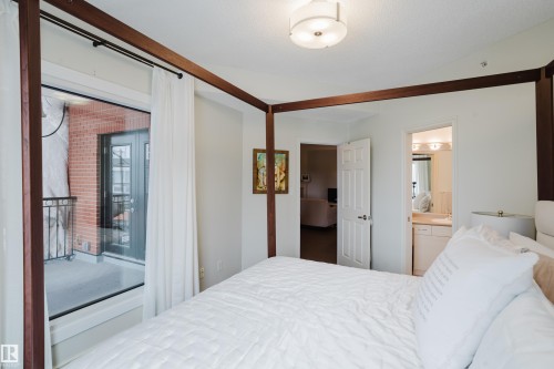 The bedroom features white walls, a ceiling-mounted light fixture, and a large glass door providing access to a balcony with a black wrought iron railing and a brick exterior wall - 206 10108 125 Street, Edmonton, AB - Indoor Photo Showing Bedroom