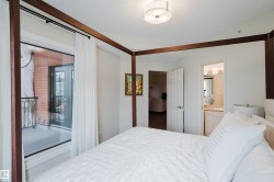 The bedroom features white walls, a ceiling-mounted light fixture, and a large glass door providing access to a balcony with a black wrought iron railing and a brick exterior wall - 