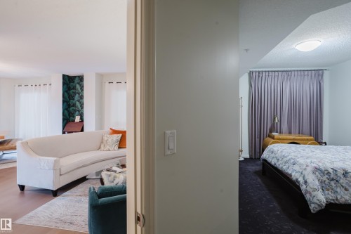 The property features a bright living area with light hardwood floors and a decorative accent wall, visible from a separate room with dark flooring and privacy curtains - 206 10108 125 Street, Edmonton, AB - Indoor Photo Showing Bedroom