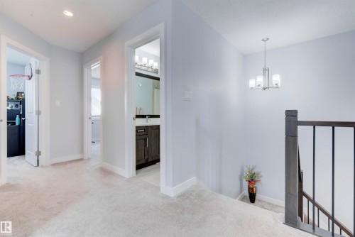 The property features a bright hallway with light-colored carpeting, a contemporary chandelier, and a dark wood staircase railing - 8452 Cushing Crest, Edmonton, AB - Indoor Photo Showing Other Room
