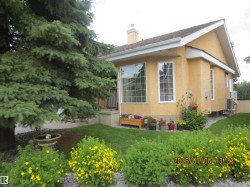 Exterior stucco siding with a pitched roof, featuring multiple windows with blinds, a manicured lawn, and mature evergreen trees - 