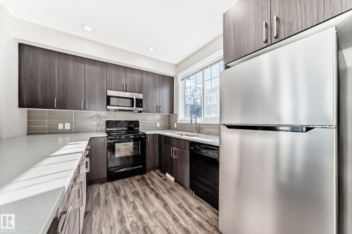 The kitchen features dark wood cabinetry, light countertops, and a full suite of stainless steel and black appliances - 75 2803 James Mowatt Trail, Edmonton, AB - Indoor Photo Showing Kitchen With Double Sink With Upgraded Kitchen