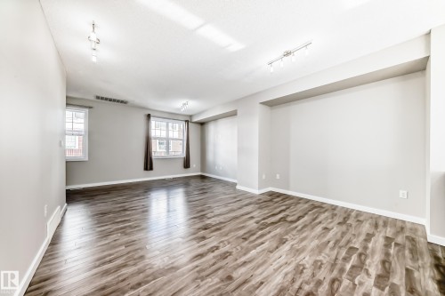 Spacious open area featuring wood-look flooring, light-colored walls, and windows with brown curtains - 75 2803 James Mowatt Trail, Edmonton, AB - Indoor Photo Showing Other Room