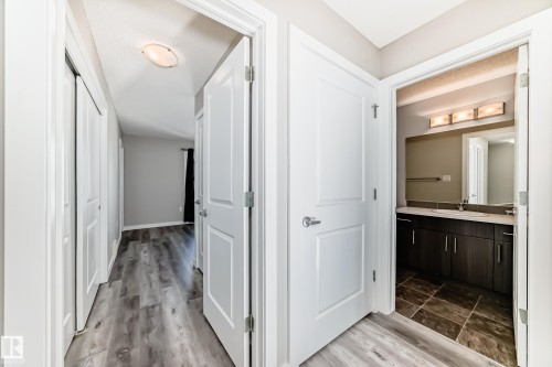 The property features hardwood style flooring in the hallway, leading to a bathroom with tiled flooring, a dark wood vanity, and a mirror with overhead lighting - 75 2803 James Mowatt Trail, Edmonton, AB - Indoor