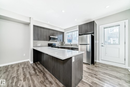 Contemporary kitchen featuring dark wood cabinetry, a light-colored island with a countertop, stainless steel appliances, and wood-look flooring - 75 2803 James Mowatt Trail, Edmonton, AB - Indoor Photo Showing Kitchen With Upgraded Kitchen