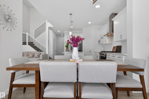 The dining area features a wooden table with light-colored chairs, open to the kitchen with white cabinetry and a stainless steel range hood, and a staircase with a dark railing - 7344 177 Avenue, Edmonton, AB - Indoor Photo Showing Dining Room
