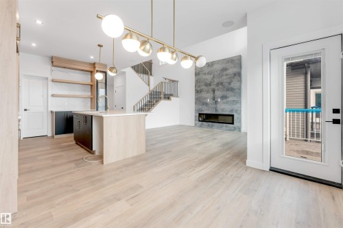 This open-concept living space features light wood flooring, a central kitchen island with a sink and pendant lighting, and a modern fireplace with a textured surround - 22 Meadowgrove Landing, Spruce Grove, AB - Indoor Photo Showing Other Room