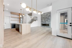 This open-concept living space features light wood flooring, a central kitchen island with a sink and pendant lighting, and a modern fireplace with a textured surround - 