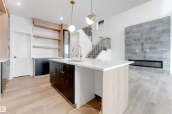 This open-concept living space features light wood-look flooring, a kitchen island with a white countertop, and a modern fireplace with a large gray tile surround - 