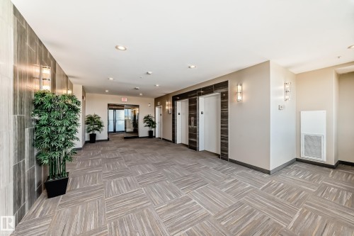 The property features a well-lit lobby with recessed lighting and wall sconces, showcasing a patterned floor, and two elevators with dark wood-toned surrounds - 506 11080 Ellerslie Road, Edmonton, AB - Indoor Photo Showing Other Room