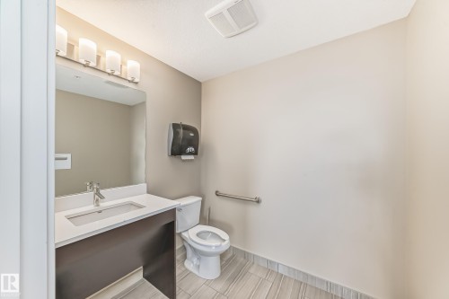 Bathroom featuring a vanity with a white countertop and an integrated sink, a toilet, a wall-mounted paper towel dispenser, and light-toned tile flooring - 506 11080 Ellerslie Road, Edmonton, AB - Indoor Photo Showing Bathroom
