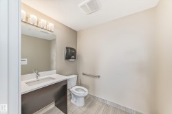 Bathroom featuring a vanity with a white countertop and an integrated sink, a toilet, a wall-mounted paper towel dispenser, and light-toned tile flooring - 