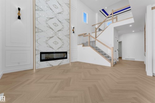 The interior features herringbone flooring, a modern linear fireplace set within a large marble-patterned wall, and a staircase with a light wood handrail and glass panels - 12 Dalquist Bay, Leduc, AB - Indoor With Fireplace