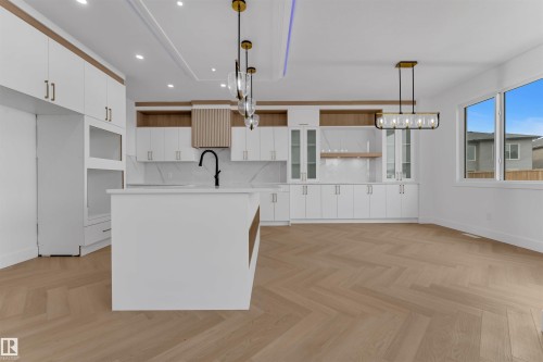This expansive kitchen features light wood flooring laid in a herringbone pattern, white cabinetry with gold-toned hardware, a central island with an integrated sink, and an elegant pendant lighting fixture - 12 Dalquist Bay, Leduc, AB - Indoor Photo Showing Kitchen