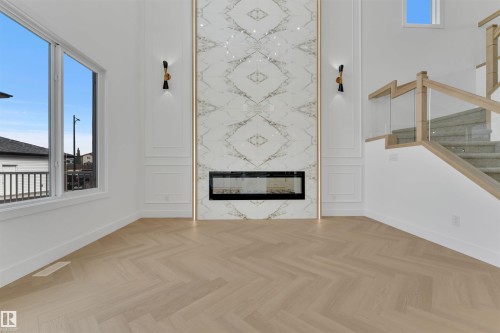 This refined space features herringbone pattern flooring, a striking fireplace with a marble-like surround, and a modern staircase with glass panel railing - 12 Dalquist Bay, Leduc, AB - Indoor With Fireplace