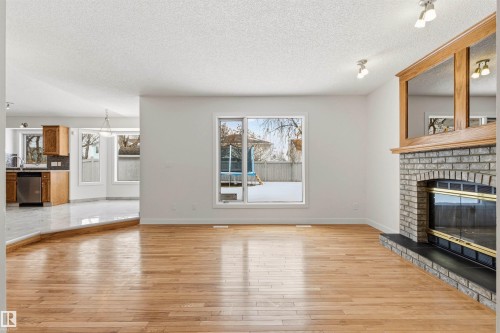 Living area featuring hardwood flooring, a brick fireplace with a wood mantle, and a large window overlooking the yard - 3668 31A Street, Edmonton, AB - Indoor Photo Showing Living Room With Fireplace