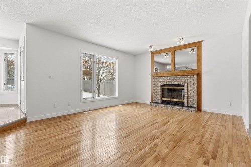 Inviting living area featuring hardwood flooring, a brick fireplace with a wood mantel and mirrored accents, and a large window providing views of the outdoors - 3668 31A Street, Edmonton, AB - Indoor Photo Showing Living Room With Fireplace