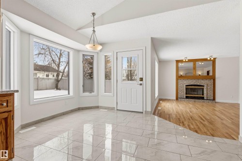 An inviting entryway with polished tile flooring and a bay window, leading to a living area featuring hardwood floors and a brick fireplace with a decorative wood mantel - 3668 31A Street, Edmonton, AB - Indoor With Fireplace