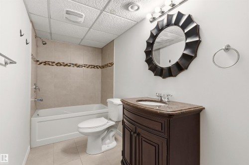 Bathroom featuring a white bathtub with tiled surround and decorative accent band, a toilet, a dark wood vanity with a light-colored countertop and undermount sink, and a decorative mirror - 3668 31A Street, Edmonton, AB - Indoor Photo Showing Bathroom