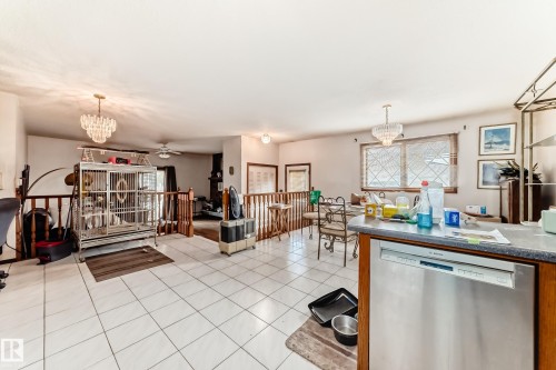 Open-concept living space with white tiled flooring, featuring a kitchen peninsula with a stainless steel dishwasher, a chandelier, and wood railings - 208 Willow Crescent, Wetaskiwin, AB - Indoor