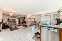 Open-concept living space with white tiled flooring, featuring a kitchen peninsula with a stainless steel dishwasher, a chandelier, and wood railings - 