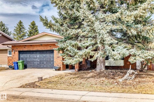 Brick exterior residence featuring a prominent mature evergreen tree, an attached two-car garage with a panel door, and a paved driveway - 208 Willow Crescent, Wetaskiwin, AB - Outdoor