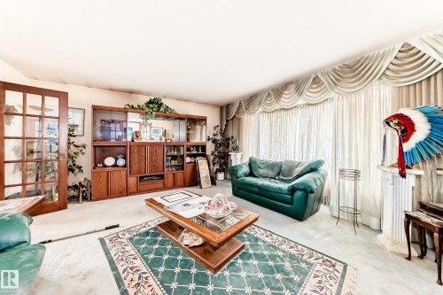 Living area featuring broad window treatments, a built-in wood-finish media console with glass display cabinets, and a paneled glass door - 208 Willow Crescent, Wetaskiwin, AB - Indoor Photo Showing Living Room