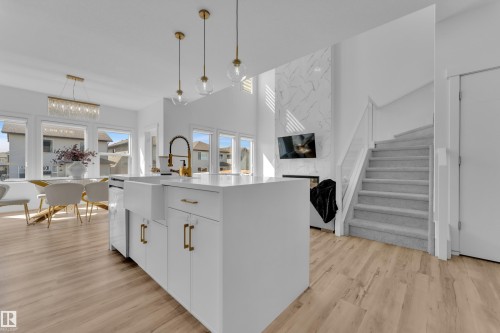 The open concept living area features light-toned flooring, a bright white kitchen island with gold-toned hardware, and a staircase with a gray carpet runner and white railings - 7815 174 Avenue, Edmonton, AB - Indoor Photo Showing Kitchen With Upgraded Kitchen