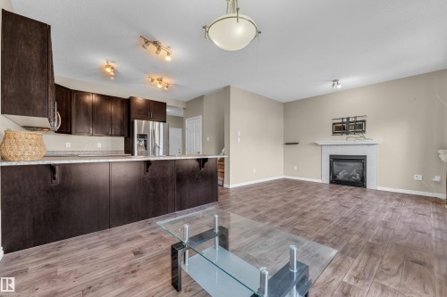 Open-concept living area featuring wood-finish flooring, a kitchen island with dark cabinetry and light countertops, stainless steel appliances, a fireplace with a white mantle, and track lighting - 2526 19A Avenue, Edmonton, AB - Indoor With Fireplace