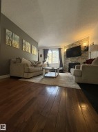 Expansive living area featuring rich wood-finish flooring, a contemporary stone-clad fireplace, and a large window with natural light - 