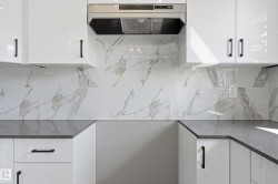 The kitchen features white cabinetry with dark hardware, a stainless steel range hood, stone countertops, and a white and grey patterned tile backsplash - 