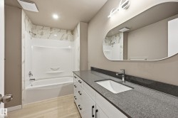 Bathroom featuring a white bathtub and shower with a tile surround, a vanity with a dark countertop and white cabinets, a rectangular undermount sink, and a modern mirror with integrated lighting - 