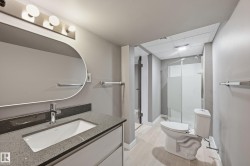 The bathroom features a modern vanity with a rectangular sink and a stone countertop, a wall-mounted mirror with overhead lighting, a toilet, and a glass-enclosed shower - 