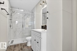 Bathroom featuring a glass-enclosed shower with marble-look wall panels, a single vanity with storage, wood-finish flooring, and a contemporary light fixture - 