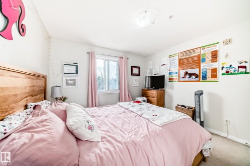 A spacious room featuring a light-colored wood headboard, a window with pink curtains, and light-colored carpeting - 317 7511 171 Street, Edmonton, AB - Indoor Photo Showing Bedroom