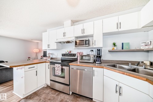 The kitchen features white cabinetry, stainless steel appliances, a double basin sink, and a floating shelf - 317 7511 171 Street, Edmonton, AB - Indoor Photo Showing Kitchen With Double Sink