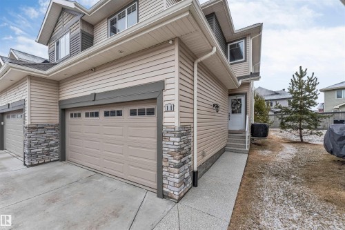 This property features a beige exterior with stone accents, a two-car garage with paneling and windows, and a concrete driveway - 11 4835 Wright Drive, Edmonton, AB - Outdoor