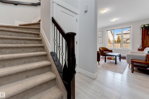 Carpeted staircase with dark wood handrail and decorative iron spindles, complemented by light wood-finish flooring and a bright living area with a large window - 11 4835 Wright Drive, Edmonton, AB - Indoor Photo Showing Other Room