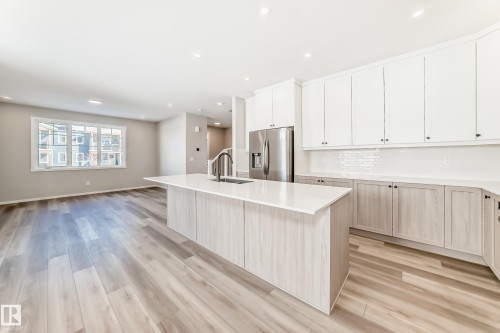 The property features a bright kitchen and living area with light wood flooring, white cabinetry, a kitchen island with a sink, and stainless steel appliances - 840 Stillwater Boulevard, Edmonton, AB - Indoor Photo Showing Kitchen With Upgraded Kitchen