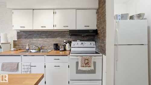 Compact kitchen featuring white cabinetry, wood-finish countertops, a dual-basin sink, and a stacked stone-look backsplash - 12 14120 80 Street, Edmonton, AB - Indoor Photo Showing Kitchen With Double Sink
