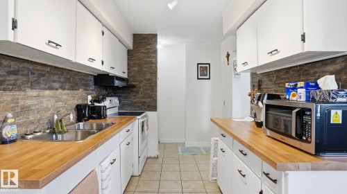 Efficient kitchen featuring white cabinetry with dark pulls, wood-finish countertops, a stainless steel sink, a stone-look tile backsplash, and ceramic floor tile - 12 14120 80 Street, Edmonton, AB - Indoor Photo Showing Kitchen With Double Sink
