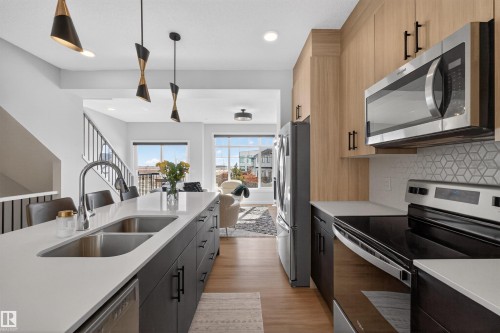 Contemporary kitchen featuring wood-tone cabinetry, stainless steel appliances, a white quartz countertop island with a double sink, and a distinctive geometric backsplash - 28 6214 Cartmell Crest, Edmonton, AB - Indoor Photo Showing Kitchen With Double Sink With Upgraded Kitchen