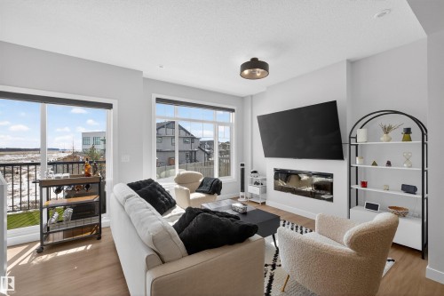The living area features light-toned flooring, large windows, and a wall-mounted fireplace - 28 6214 Cartmell Crest, Edmonton, AB - Indoor Photo Showing Living Room With Fireplace