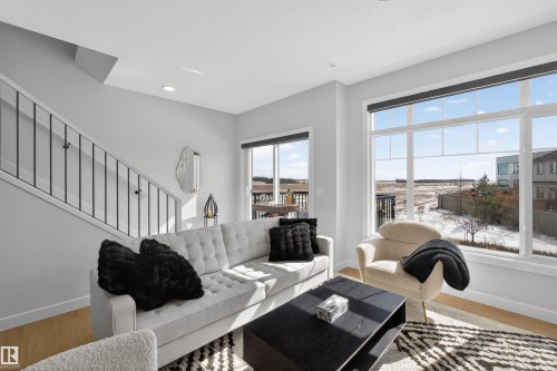 The living area features large windows providing views of the surrounding landscape, light-toned walls, and a staircase with contemporary black railings - 28 6214 Cartmell Crest, Edmonton, AB - Indoor Photo Showing Living Room